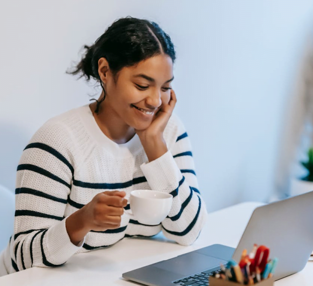 Une fille souriante devant son ordinateur en train de boire un café et de suivre une conversation vidéo.